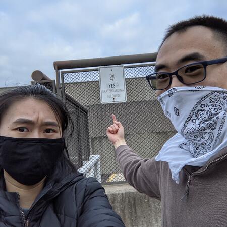 A man and woman, wearing face masks, look into the camera as the man points to an outdoor fence sign that reads, yes skateboarding allowed.