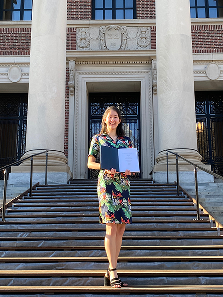 Isabel Bernhard smiling and holding her completed thesis in front of Widener Library