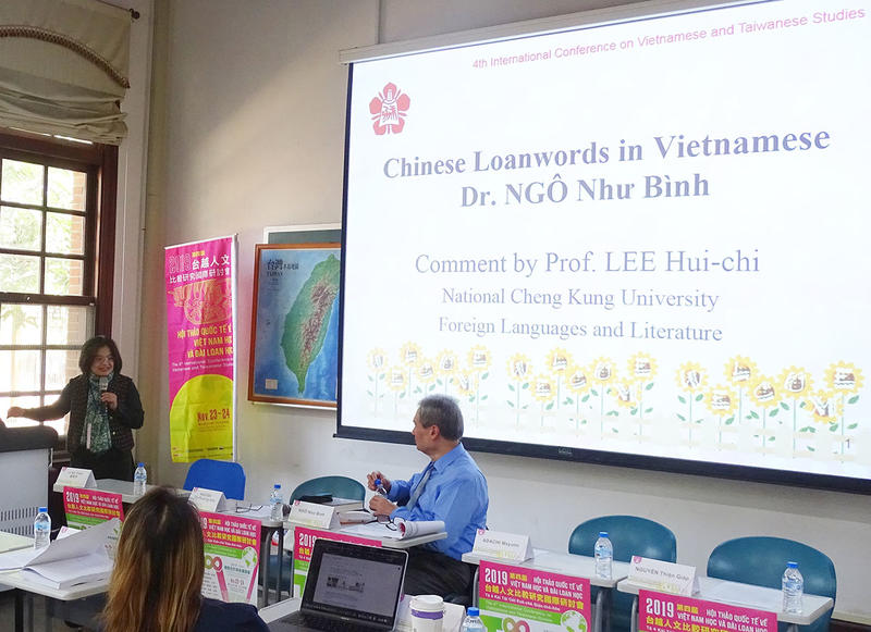 Dr. Ngo, seated in front of a projector screen bearing the title slide of his lecture on Chinese loanwords in Vietnamese, engages in conversation with a female colleague who stands at the podium speaking animatedly into a microphone.
