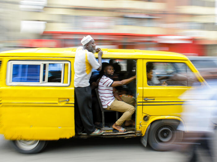 A bright yellow minibus (danfo) whizzes past, with the side van door open and two men hanging on.