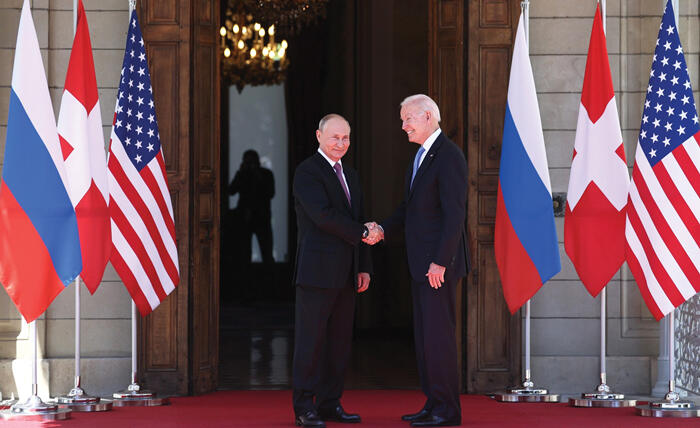 US President Joe Biden shakes hands with Russian President Vladimir Putin in front of a door flanked by both countries' flags