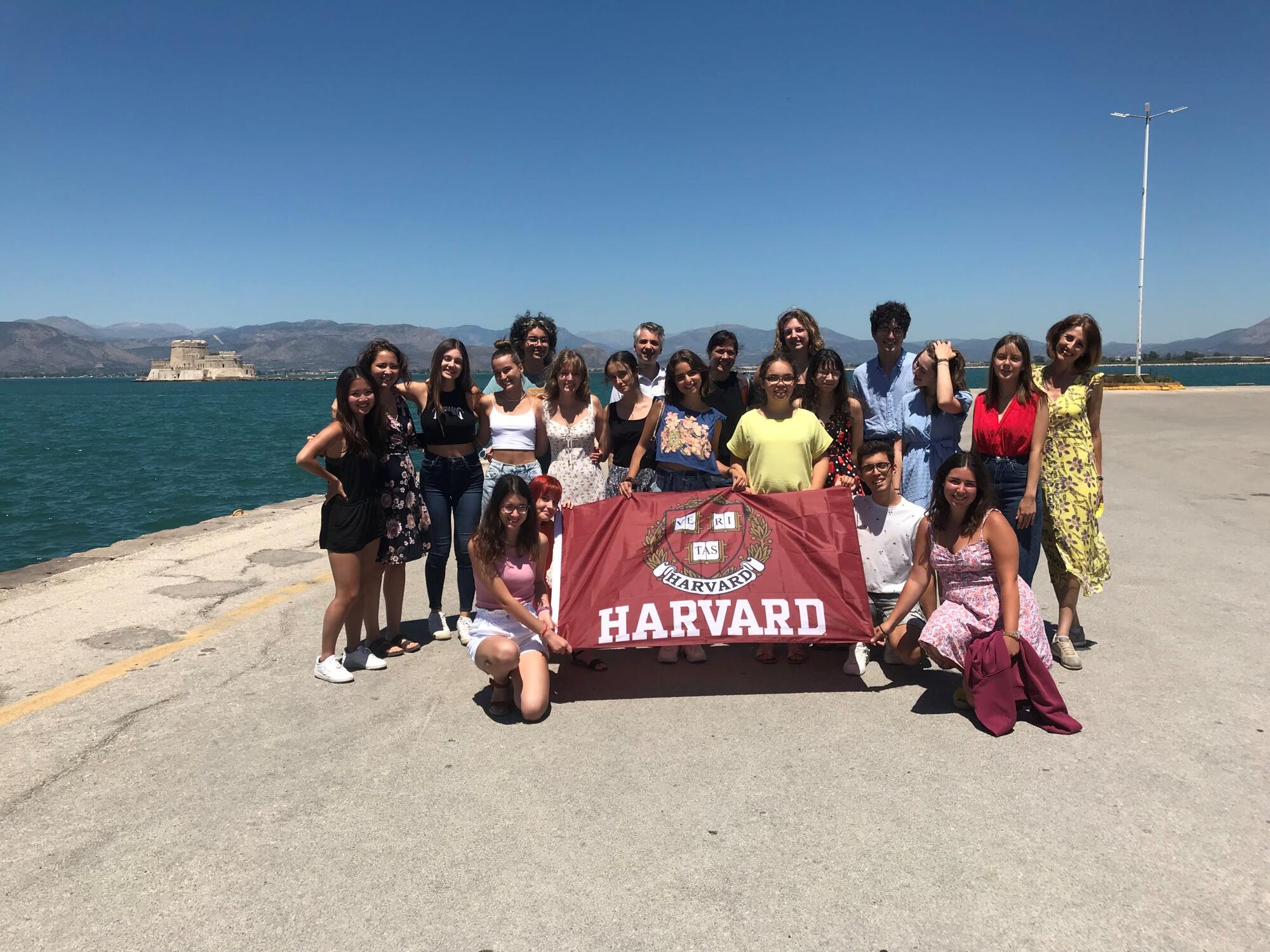 HSSP 2022 Group in front of the Port of Nafplio holding a Harvard flag.