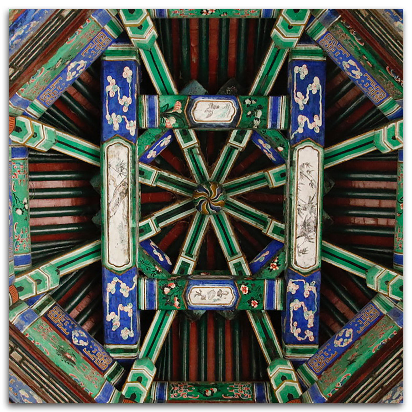 An intricate pattern of wooden ceiling beams in the shape of a dharma wheel, seen from below, painted in blue green red and white