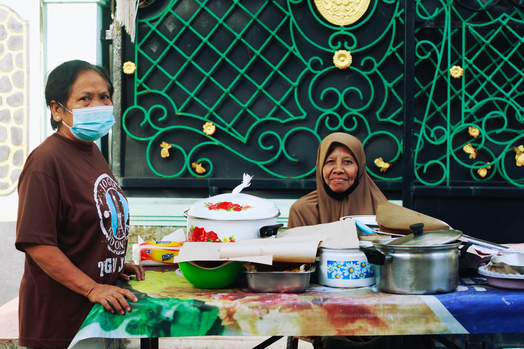 Two Indonesian women at the camera. One is sitting behind a market table, wearing an abaya and a surgical mask pulled under her chin; the other is standing next to the table, wearing a graphic tee and with a surgical mask over her face.