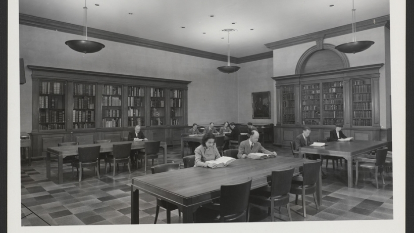 View of the Treasure Room, Langdell Hall with men and women sitting at tables. Bookcase pediment is blank, prior to the addition of the painted shield.
