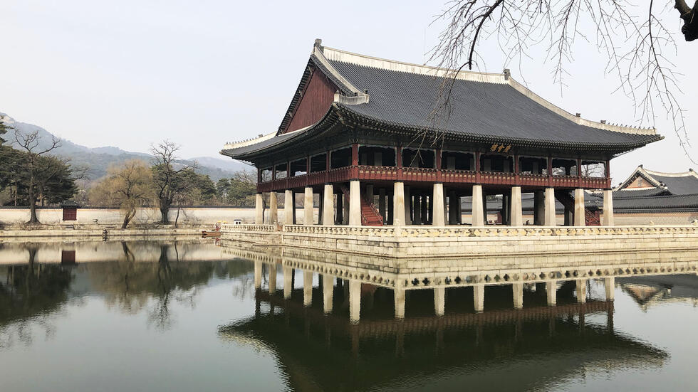 Inside an expansive Korean palace complex, a pagoda-like structure stands on stilts in the middle of a manmade pond.
