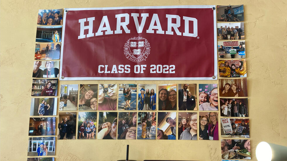 An open laptop and other supplies sits on a desk facing a textured yellow wall that is decorated with a Harvard felt banner, Class of 2022, and a collage of color photos neatly arranged in rows, displaying young people and places.