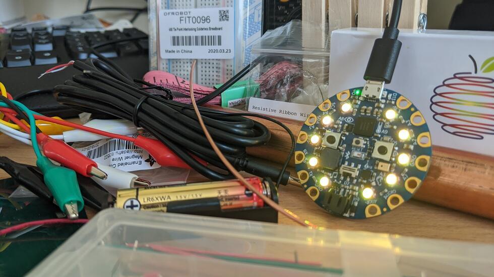 Image shows a Bluefruit device with illuminated LEDs, batteries, assorted cables, a copper pipe, a container of jumper cables, resistive wire, and a small breadboard.