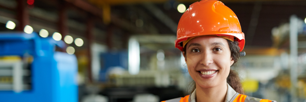 A young woman wearing a safety vest and hard hat smiles in a warehouse