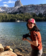Photo of Julie smiling in front of a lake and mountain, holding a camera