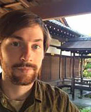 Headshot of Daniel Borengasser, showing a young man with a neutral expression, short red-brown hair, and facial hair in front of a Japanese-style garden.