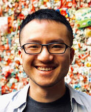 A man with short black hair and glasses smiles broadly, standing in front of a multicolored wall