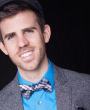 Smiling man in a blue shirt and grey wool jacket, with a plaid bow tie